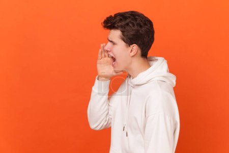 Photo for Side view portrait of angry or shocked handsome brunette young man wearing white hoodie standing, looking forward and screaming. Indoor studio shot isolated on orange background. - Royalty Free Image