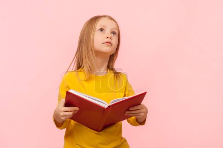 Portrait of dreaming blonde little girl holding book, looking away, dreaming, do not want to study, thinking, wearing yellow jumper. Indoor studio shot isolated on pink background.