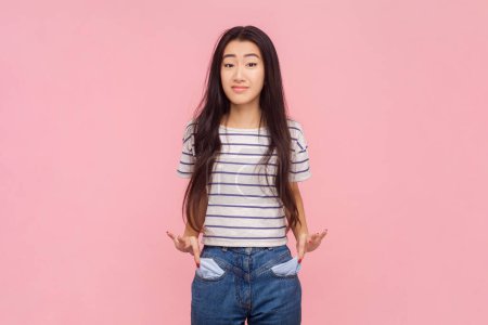 Portrait of poor displeased woman with long brunette hair showing her empty pockets expressing negative emotions, wearing striped T-shirt. Indoor studio shot isolated on pink background.