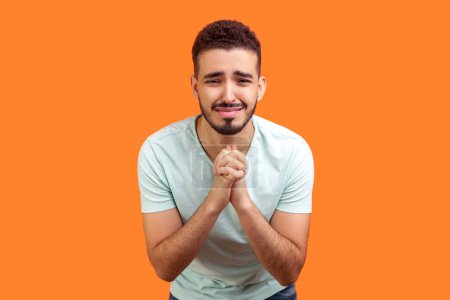 Portrait of sad crying young bearded man wearing T-shirt keeps hands together pleading praying apology. Indoor studio shot isolated on orange background.