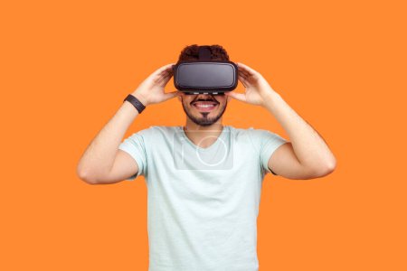 Téléchargez les photos : Portrait d'un jeune homme barbu souriant portant un T-shirt debout avec un casque de réalité virtuelle regardant une vidéo intéressante. Studio intérieur tourné isolé sur fond orange. - en image libre de droit