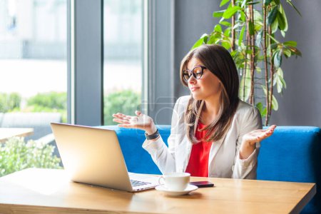 Photo for Portrait of puzzled confused woman working on laptop having online internet meeting shrugging shoulders, wearing jacket and re shirt. Indoor shot, cafe or office background. - Royalty Free Image