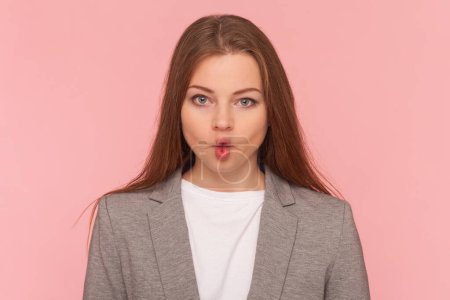 Portrait of childish playful woman with brown hair making fish lips, looking at camera, having fun, grimacing, wearing business suit. Indoor studio shot isolated on pink background.
