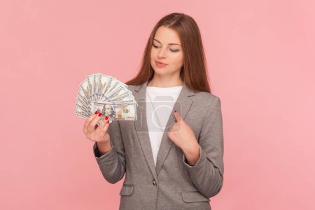 Photo for Portrait of proud rich woman with brown hair holding dollars banknotes, pointing at herself, earning big sum of money, wearing business suit. Indoor studio shot isolated on pink background. - Royalty Free Image