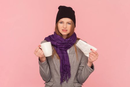 Portrait of ill sick woman with brown hair having grippe symptoms, holding napkin and cup in hands, drinking tea, wearing hat and scarf. Indoor studio shot isolated on pink background.