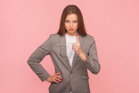 Portrait d'une enseignante sérieuse et stricte aux cheveux bruns debout avec la main sur la hanche et levant le doigt vers le haut, avertissant, portant un costume d'affaires. Studio intérieur tourné isolé sur fond rose.