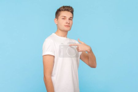 Portrait of egoistic confident man wearing white t-shirt standing pointing at himself, being proud of hos achievements, looking at camera. Indoor studio shot isolated on blue background