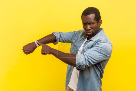 Photo for Portrait of serious bossy man standing pointing at his smartwatch looking at camera with irritated face, wearing denim casual shirt. Indoor studio shot isolated on yellow background. - Royalty Free Image
