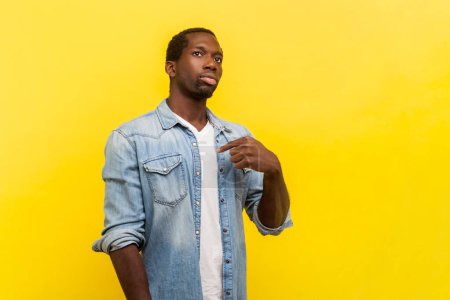 Portrait of proud successful satisfied man pointing at himself looking at camera with egoistic facial expression, wearing denim casual shirt. Indoor studio shot isolated on yellow background.