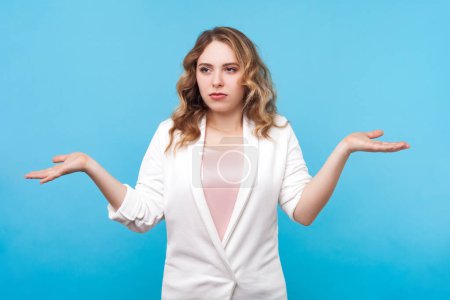 Photo for Portrait of puzzled confused blond woman with wavy hair standing shrugging shoulders not sure looks helpless, wearing white shirt. Indoor studio shot isolated on blue background. - Royalty Free Image