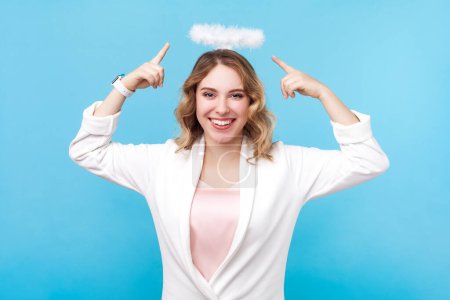 Portrait of smiling angelic blond woman with wavy hair pointing at nimb over her hair looking at camera with happy expression, wearing white shirt. Indoor studio shot isolated on blue background.