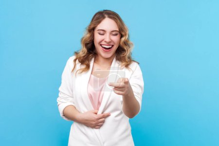 Portrait of funny laughing blond woman with wavy hair holding belly pointing to camera hearing joke, choosing, wearing white shirt. Indoor studio shot isolated on blue background.