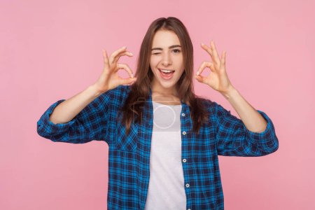 Photo for Portrait of playful funny brown haired woman showing ok gestures winking to camera, having satisfied expression, wearing checkered shirt. Indoor studio shot isolated on pink background. - Royalty Free Image
