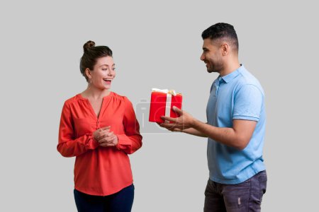 Photo for Portrait of happy positive couple standing together man giving present box to his girlfriend celebrating birthday anniversary. Indoor studio shot isolated on gray background. - Royalty Free Image