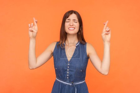 Photo for Portrait of hopeful brunette woman wearing denim dress crossing fingers closed eyes praying for good luck waiting for miracle. Indoor studio shot isolated on orange background. - Royalty Free Image