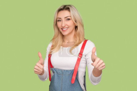 Téléchargez les photos : Portrait of smiling delighted adult blond woman showing thumbs up, like gesture, positive feedback, wearing denim overalls. Indoor studio shot isolated on light green background - en image libre de droit