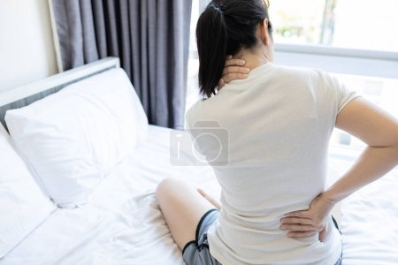 Foto de Mujer asiática adulta tiene dolor en el cuello, hombro y dolor de espalda sentado en la cama en el dormitorio, personas estresadas que sufren de dolor de espalda, tensión y lesión en su nuca en la almohada incómoda y colchón - Imagen libre de derechos