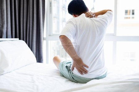 Téléchargez les photos : Asiatique homme adulte ont des douleurs dans le cou, les épaules et le dos assis sur le lit dans la chambre, les personnes stressées souffrant de maux de dos, de tension et de blessures à la nuque sur un oreiller et un matelas inconfortables - en image libre de droit