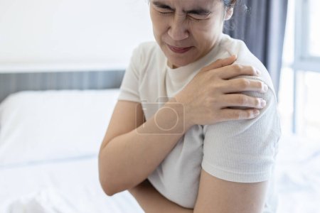 Foto de Mujer asiática de mediana edad que sufre de dolor en el hombro, dislocación del hombro, entumecimiento y debilidad dolorosa, paciente mujer cansada con articulación dislocada del hombro, malestar causado por enfermedad o lesión - Imagen libre de derechos