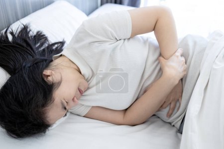 Femme asiatique souffrant de douleurs abdominales, couchée sur le lit, gastrite, ulcère gastrique, patiente souffrant de maux d'estomac, symptôme de troubles gastro-intestinaux, ulcère d'estomac, problème gastrique, soins de santé