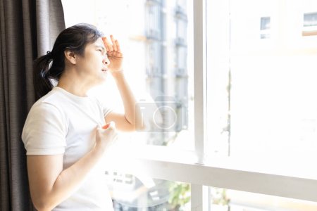 Téléchargez les photos : Femme asiatique regardant par la fenêtre à la lumière du soleil forte, temps chaud, vague de chaleur à haute température, rayons ultraviolets sévères du soleil, risque de dommages aux yeux, coup de soleil, réchauffement climatique, changement climatique, El Nino, soins de santé - en image libre de droit