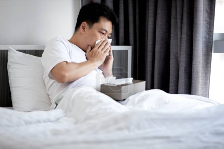 Hombre de mediana edad tiene una nariz congestionada, secreción nasal, golpe o limpiar la nariz con papel tisú, asiático masculino tiene fiebre y resfriado o gripe, enfermedad respiratoria, infección del tracto respiratorio, descansando en la cama en casa