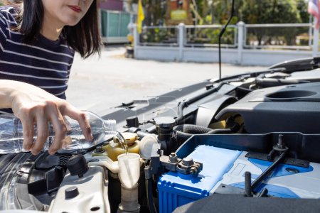 Asian woman pouring clean water from bottle into the windshield washer fluid tank of a car,female filling the windshield washer fluid,check liquid level in reservoir at engine room,maintenance concept