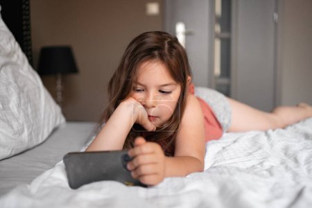Chica de 5 años viendo dibujos animados en el teléfono móvil en la cama. interior de lujo real. Foto de alta calidad