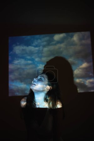 Téléchargez les photos : Jeune adulte belle femme z-gen avec des nuages image projetée sur elle avec projecteur. Photo de haute qualité - en image libre de droit