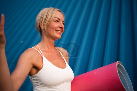 Foto de Mujer hermosa de mediana edad con tapete deportivo al aire libre sobre fondo de pared azul. Hace calor. Foto de alta calidad - Imagen libre de derechos