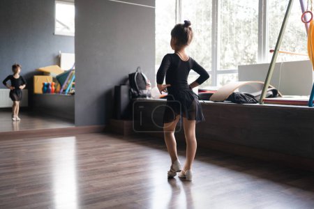 Téléchargez les photos : Enfant fille debout en costume de sport noir en studio de danse pendant la posture d'entraînement. 4 5 ans d'âge préscolaire. Développement physique sain. Photo de haute qualité - en image libre de droit