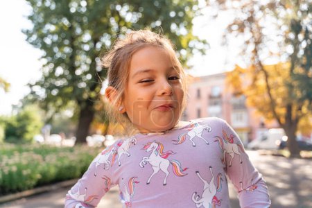 Téléchargez les photos : Portrait d'une jeune fille caucasienne drôle de 5-6 ans faisant un visage à l'extérieur dans une ville. Portrait en gros plan d'un enfant grimaçant. Photo de haute qualité - en image libre de droit