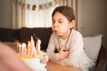 Un enfant ferme les yeux pour faire un v?u d'anniversaire, soufflant des bougies sur un gâteau à la licorne, entouré d'une joyeuse ambiance de fête à la maison. Photo de haute qualité