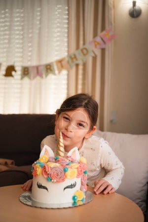 A young girl poses with a gleeful smile beside her magical unicorn birthday cake, ready to celebrate her special day with sweetness and joy. High quality photo