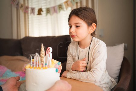 Ein Kind schließt die Augen, um einen Geburtstagswunsch zu formulieren, bläst Kerzen auf eine Einhorntorte, umgeben von fröhlicher Feierstimmung zu Hause. Hochwertiges Foto