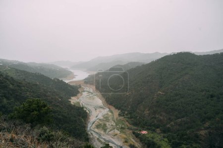A tranquil scene of a winding river cutting through a lush valley, with fog blanketing the distant hills and a solitary house visible. High quality photo