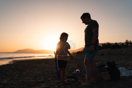 Ein Erwachsener und ein Kind unterhalten sich vor malerischer Strandkulisse, während die untergehende Sonne die Szenerie in weiches, bernsteinfarbenes Licht taucht. Hochwertiges Foto