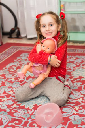 Photo for A little girl in red is played with a doll. Swaddles the doll at home on the carpet - Royalty Free Image