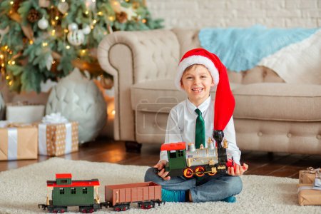 Foto de Niño feliz con un regalo, tren de juguete, bajo el árbol de Navidad en una mañana de Año Nuevo. Un tiempo de milagros y realización de deseos. Feliz Navidad.. - Imagen libre de derechos