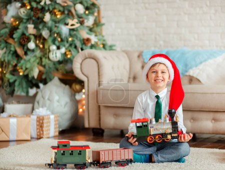Foto de Niño feliz con un regalo, tren de juguete, bajo el árbol de Navidad en una mañana de Año Nuevo. Un tiempo de milagros y realización de deseos. Feliz Navidad.. - Imagen libre de derechos