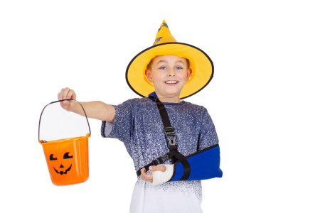 Niño en un sombrero de mago y capa brillante, sonriendo con un cubo de Halloween, listo para truco o trato