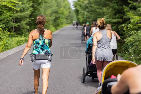 A la izquierda hay un instructor que da instrucciones aeróbicas y cardiovasculares a un grupo de madres nuevas que caminan con sus bebés en cochecitos