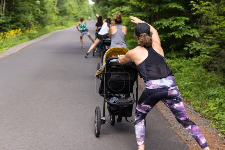 Téléchargez les photos : Groupe de mères faisant de l'exercice cardio ensemble sur une route asphaltée avec de la verdure sur les côtés avec leurs bébés dans des poussettes devant eux - en image libre de droit