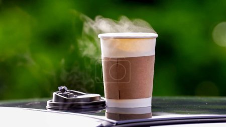 Coffee takeaway in a paper cup on top of the car roof green tree background at sunrise in the morning,  selective focus, soft focus.