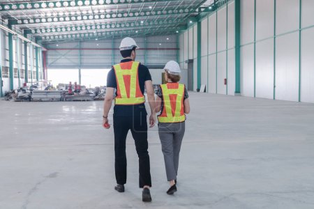 Foto de Ingenieros masculinos y femeninos están caminando en el almacén dentro de una fábrica industrial. Los ingenieros usan sombreros duros y chalecos reflectantes para mayor seguridad. - Imagen libre de derechos