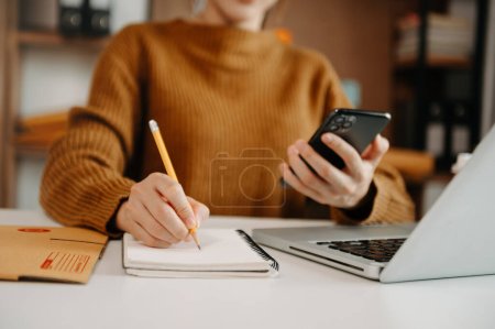 Photo for Partial view of woman working in office at table with mobile phone, laptop and making notes on paper with pencil - Royalty Free Image