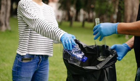 Foto de Two people keeping garbage plastic bottle into black bag at park.Clearing, pollution, ecology and plastic concept. - Imagen libre de derechos