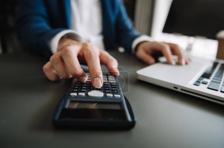 Foto de Man counting  on calculator taking from the piggy bank. hands working on calculator to calculate on desk about cost at office - Imagen libre de derechos