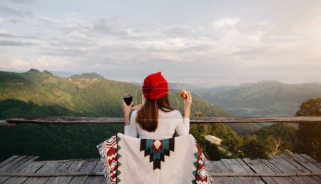 Foto de Rear view image of a female traveler sitting and holding breakfast looking at  beautiful mountain and nature view in sunlight - Imagen libre de derechos
