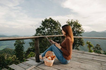 Photo for Rear view image of a female traveler sitting and holding tablet at a beautiful mountain and nature view - Royalty Free Image